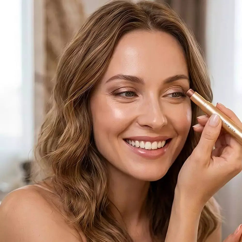 A close-up of a woman smiling while applying a makeup pencil to her eye, shown in a bright indoor setting.