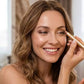 A close-up of a woman smiling while applying a makeup pencil to her eye, shown in a bright indoor setting.