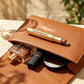 A tan faux-leather makeup bag with a gold stick eyeshadow pen resting on top, alongside a perfume bottle, lipstick, and compact mirror, set on a light stone surface with soft outdoor shadows.