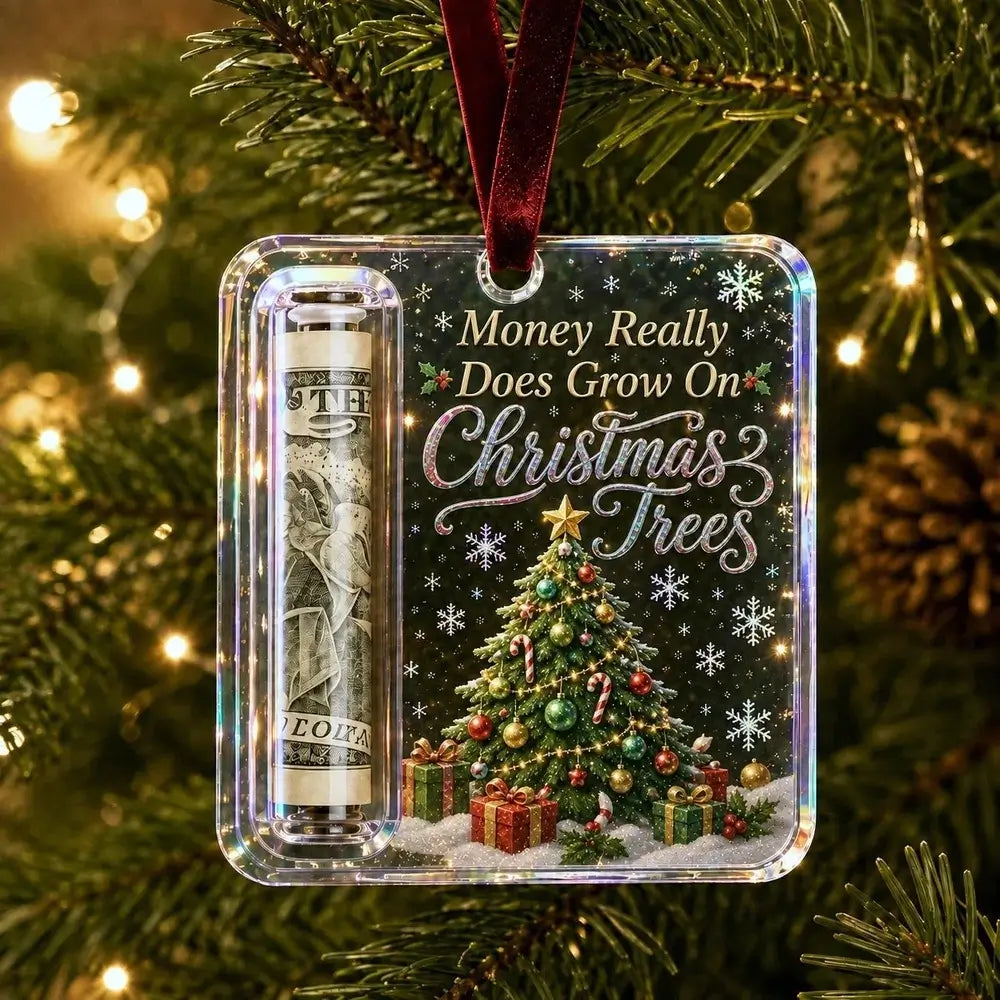 A clear acrylic Christmas ornament hanging from a green pine tree with warm string lights. The ornament contains a rolled dollar bill in a clear tube on the left and a printed Christmas tree scene with the phrase 'Money Really Does Grow On Christmas Trees' on the right.