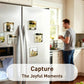 A white stainless steel refrigerator in a kitchen setting, decorated with several square clear magnets holding printed photos. A blurred man stands in the background near a window.