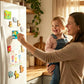 A woman holding a young child in a bright kitchen, smiling as she places a colorful refrigerator magnet on a white fridge. The fridge is decorated with various animal and family-themed magnets.