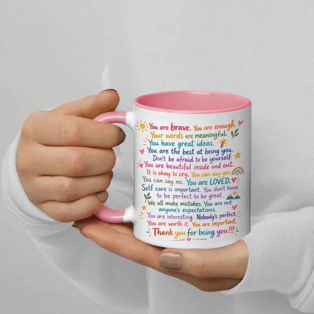 A hand holding a white ceramic mug with a pink handle and pink rim. The mug is printed with a colorful, supportive message for a child in a neat, readable font, decorated with small illustrations of suns, flowers, hearts, and rainbows.