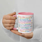 A hand holding a white ceramic mug with a pink handle and pink rim. The mug is printed with a colorful, supportive message for a child in a neat, readable font, decorated with small illustrations of suns, flowers, hearts, and rainbows.