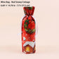 A festive red wine bag with a printed 'Red Snowy Cottage' design, featuring a green wreath, snowflakes, and a small snowy cabin. The bag is tied with a white ribbon and is shown against a solid pale pink background with a text overlay at the top detailing its dimensions.