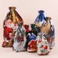 A set of Christmas-themed drawstring gift bags with various festive prints including Santa Claus, reindeer, a snowy house, and a sleigh, arranged together on a solid light beige background.