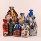 A set of Christmas-themed drawstring gift bags in various sizes and designs, including festive prints of Santa Claus, a Christmas tree, a sleigh, and holiday scenes, arranged on a solid light beige background.