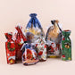 Six Christmas-themed drawstring gift bags in assorted festive colors and patterns, including Santa Claus, snowman, reindeer, snowflake, and holiday village designs, arranged on a light beige background.