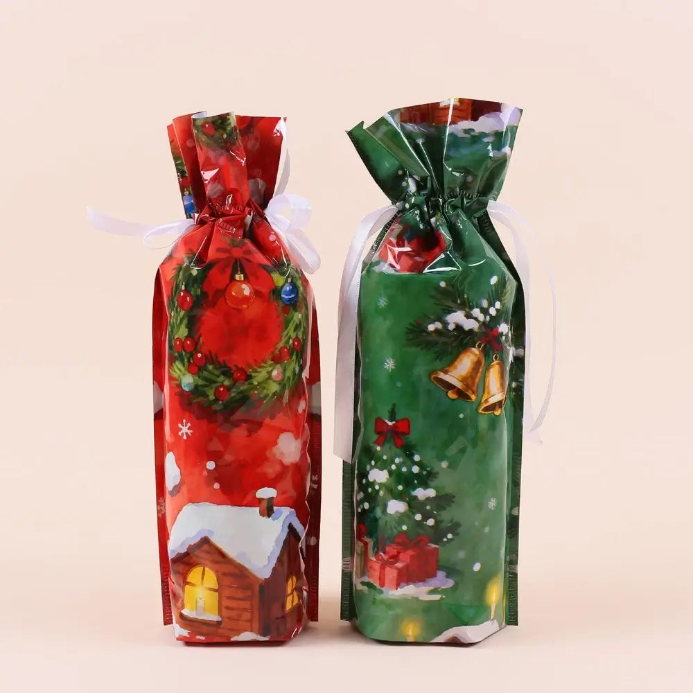 Two Christmas-themed gift pouches or bottle wraps standing upright on a light beige background. The left one is red with a holiday wreath, ornaments, snow, and a small cabin illustration, tied with a white ribbon.