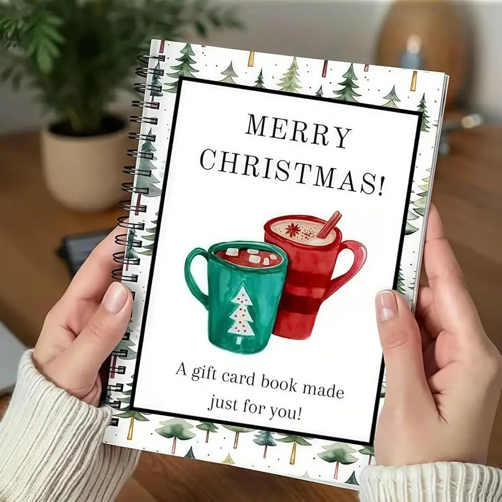 A person holds a spiral-bound gift card book with a festive Christmas cover design. The cover features the text 'MERRY CHRISTMAS! A gift card book made just for you!' above an illustration of two mugs, one teal and one red, with a Christmas tree on the teal mug.
