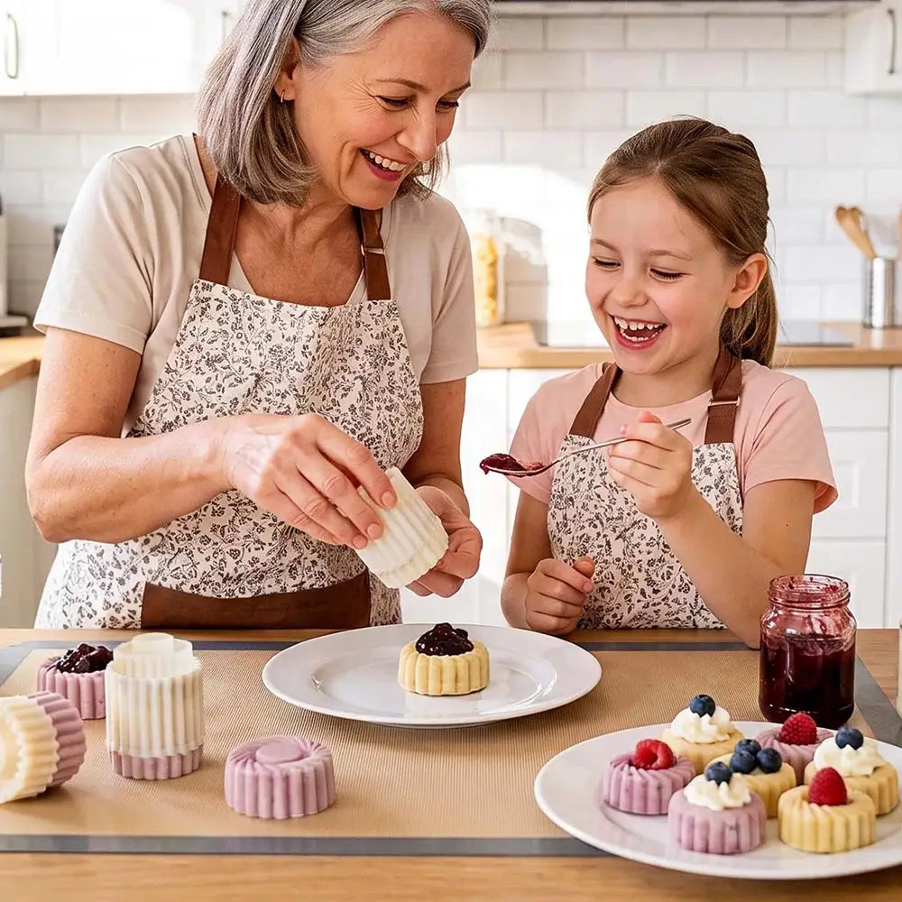 An older woman and a young girl wearing matching floral aprons smile and laugh in a bright kitchen while making small pastries. Pastries are arranged on plates on the table, including a tart with jam and several lavender and pink molded pastries.