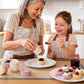 An older woman and a young girl wearing matching floral aprons smile and laugh in a bright kitchen while making small pastries. Pastries are arranged on plates on the table, including a tart with jam and several lavender and pink molded pastries.