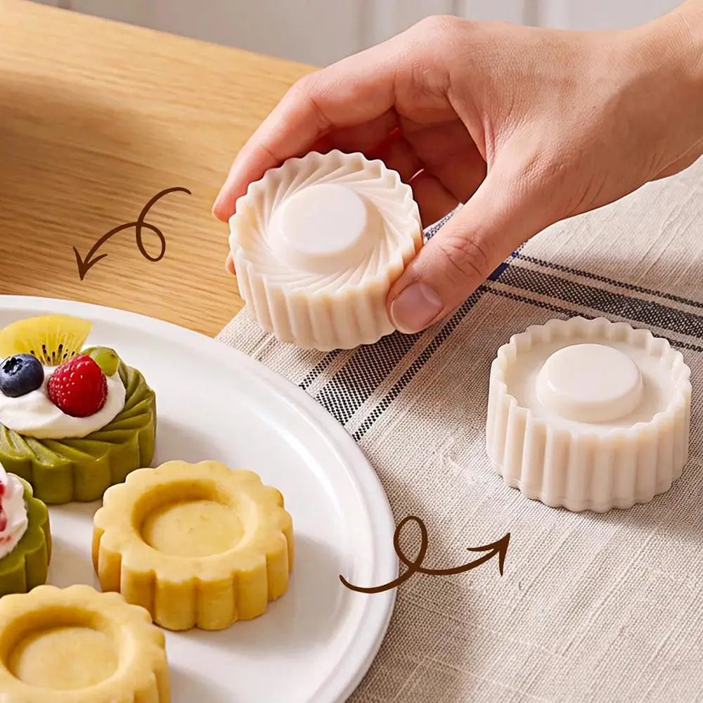 A hand holds a round white pastry cutter. The scene is set on a wooden table and a striped cloth.