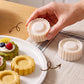 A hand holds a round white pastry cutter. The scene is set on a wooden table and a striped cloth.