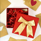 A red square gift box with a large gold satin bow on the lid, shown open with red shredded paper filler inside. Gold envelopes are scattered around on a white background.