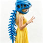 A woman wearing a blue dinosaur plush head and matching blue dinosaur back accessory, posed in a yellow dress against a plain white background.
