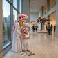 A woman and a child wearing pink dinosaur costume headpieces and a matching pink dinosaur costume dress stand in a modern museum gallery, pointing toward a large dinosaur skeleton exhibit in the background.