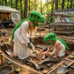 Two children wearing green dinosaur skull masks and white dresses explore an outdoor excavation site with bones and tools, with a dinosaur skeleton display and forest background.