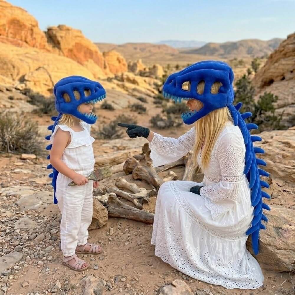 A woman and a child wearing matching blue dinosaur costume headsets and spiky back accessories pose outdoors in a rocky desert landscape. The woman wears a long white dress and black gloves, while the child wears a white sleeveless outfit.