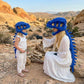 A woman and a child wearing matching blue dinosaur costume headsets and spiky back accessories pose outdoors in a rocky desert landscape. The woman wears a long white dress and black gloves, while the child wears a white sleeveless outfit.