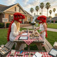 A woman and a child wearing red dinosaur helmets sit at a picnic table outdoors in a suburban backyard, surrounded by toy dinosaurs, tools, and a red-and-white checkered tablecloth. A house, lawn, and palm trees are visible in the background.
