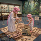 A woman and a child wearing pink dinosaur helmets and white outfits explore a paleontology exhibit inside a museum. The woman is handling a dinosaur skeleton while the child examines a ammonite shell on a display box.