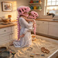 A woman and child wearing pink dinosaur skeleton head masks play in a sandbox filled with sand toys inside a cozy kitchen setting.