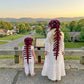 Two people standing outdoors at a scenic overlook, seen from behind, wearing matching long white dresses and white pants with dark burgundy bone-shaped headpieces that resemble dinosaur or prehistoric skull elements. A town and hills are visible in the background.