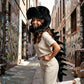 A woman wearing a black dinosaur skull and spine costume stands in a graffiti-covered alleyway, smiling and posing with one leg extended.
