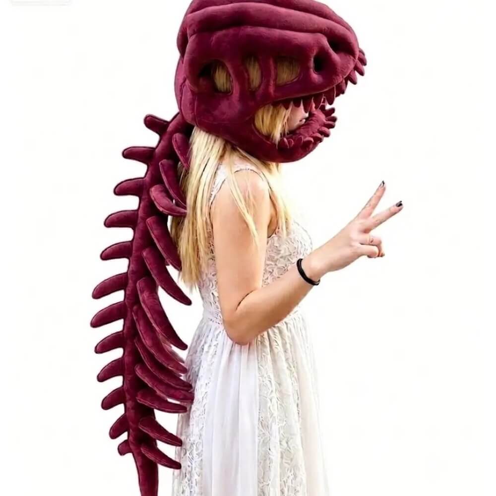 A woman wearing a large plush dinosaur skull headpiece with a long dark red tail spine, posed against a plain white background.