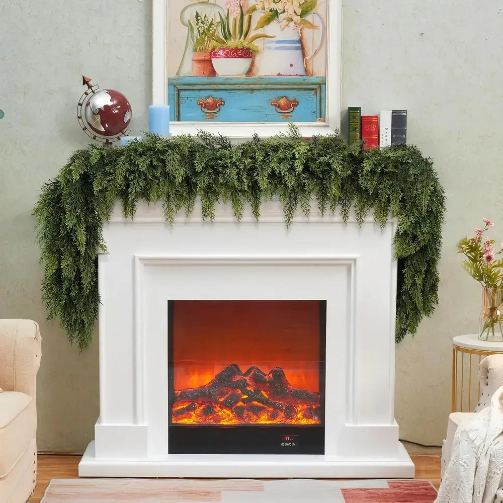 A white electric fireplace mantel with a glowing orange and red faux fire display, decorated with green artificial ferns and styled with books, a globe, and a framed picture above.