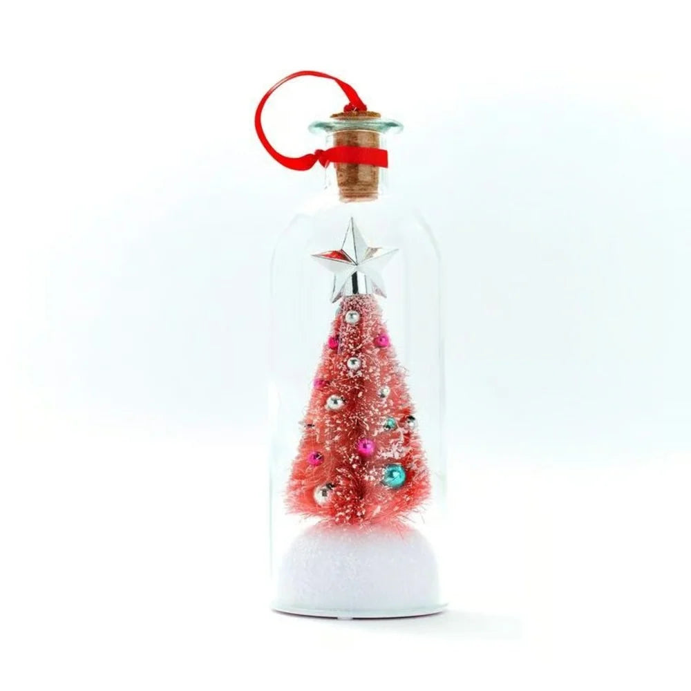 A miniature pink Christmas tree ornament inside a clear glass bottle with a cork stopper, red hanging ribbon, and white glittery base, shown on a white background.