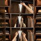A white triangular multi-hanger closet organizer shown in three stacked panels being hung on a wardrobe rod inside a closet. A person's hand and arms are visible holding the hanger, and the closet shelves around it contain folded clothes, bags, and hats.