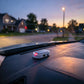 A small white rectangular electronic device with a black top panel and a glowing red light, placed on a car dashboard in front of a blurred street scene at dusk.