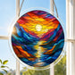 A round acrylic suncatcher with a colorful mountain river sunset and sun reflected on water hanging from a chain in front of a bright white window