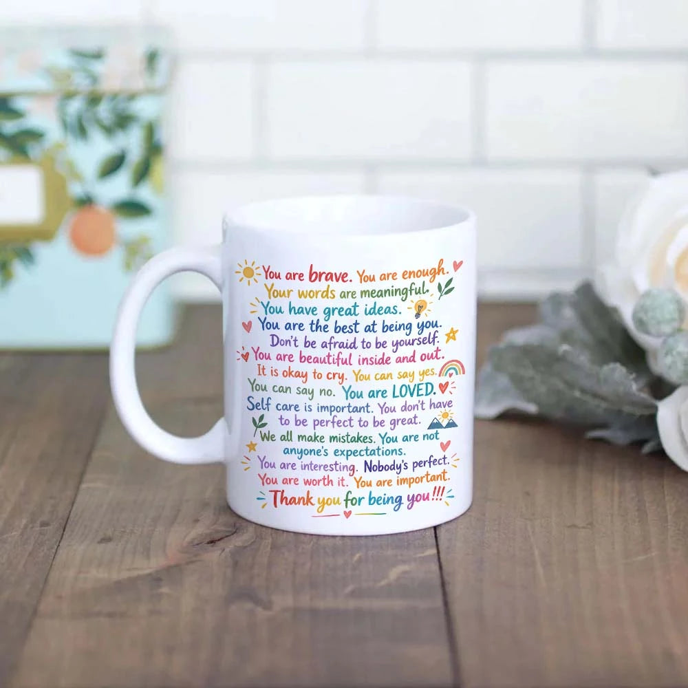 A white ceramic mug with colorful positive self-care quotes printed on it.