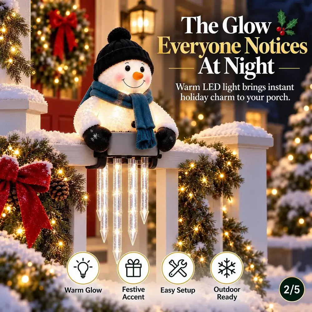 A plush snowman holiday light decoration sits on a snowy white porch railing decorated with pine garlands and warm string lights. The snowman wears a black hat and blue scarf and has glowing icicle accents hanging below.