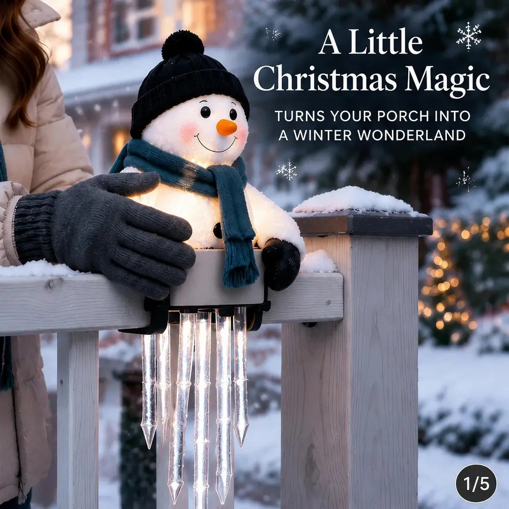 A festive snowman porch light with a black hat, blue scarf, and glowing warm face, mounted on a wooden fence in a snowy outdoor setting. A person wearing gray gloves is partially visible on the left.