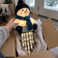 A person's hand is holding a fluffy snowman holiday light ornament inside an open cardboard box. The snowman wears a black knit hat and blue scarf, with glowing icicle lights hanging below.