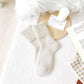 A pair of ribbed ankle socks in a soft off-white/cream color, styled in a cozy flat-lay arrangement with a wooden tray, white candle, card reading 'EVERYTHINGS', an open book, and a white fluffy rug on a light wood floor.