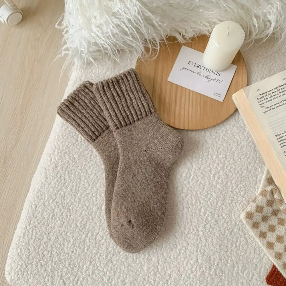 A pair of ribbed ankle socks in a warm taupe-brown color, styled in a cozy flat-lay arrangement with a wooden tray, white candle, book, and fluffy white rug.