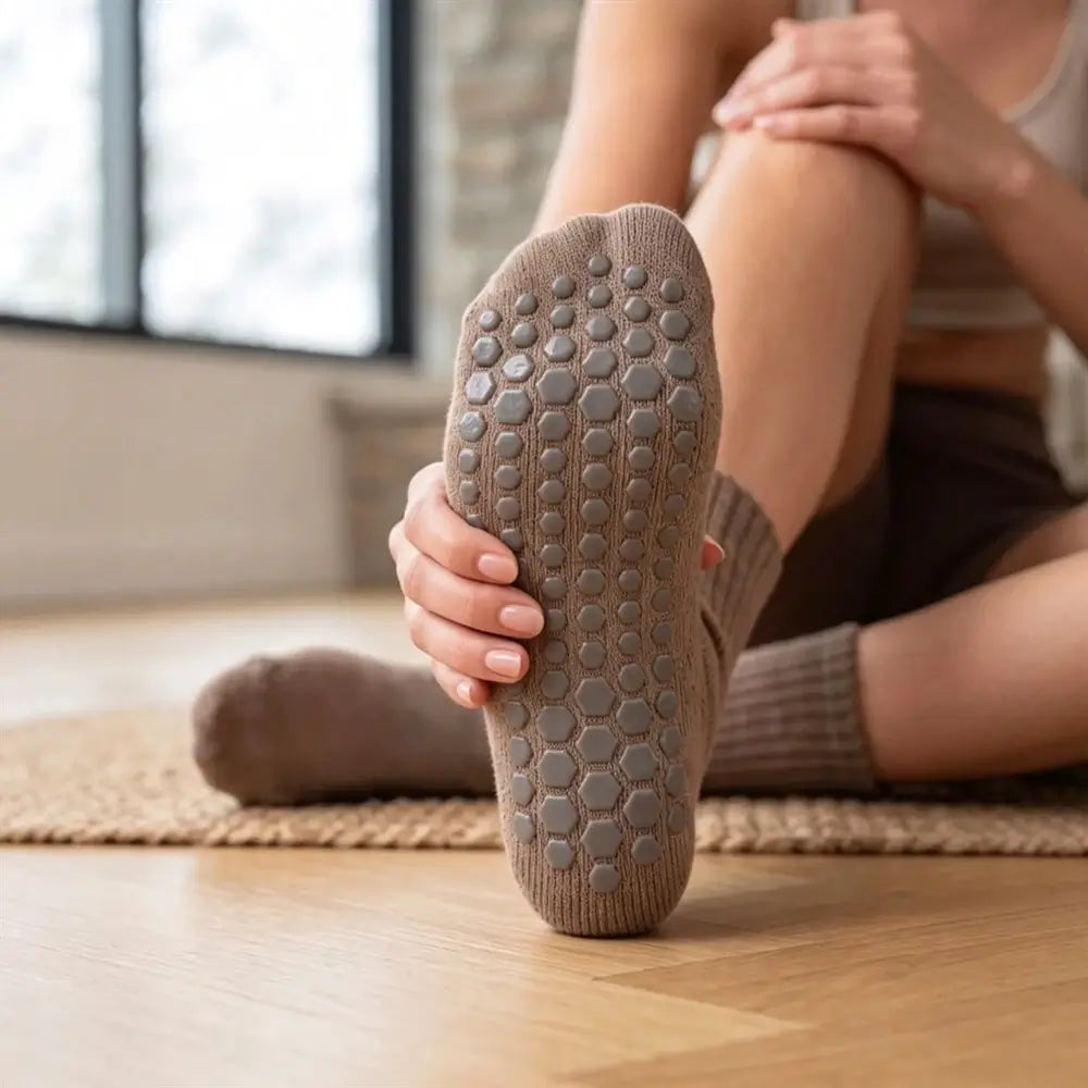 A person sitting on a wooden floor indoors, holding up one sock to show the textured non-slip sole. The sock is taupe brown with raised circular grip pads.