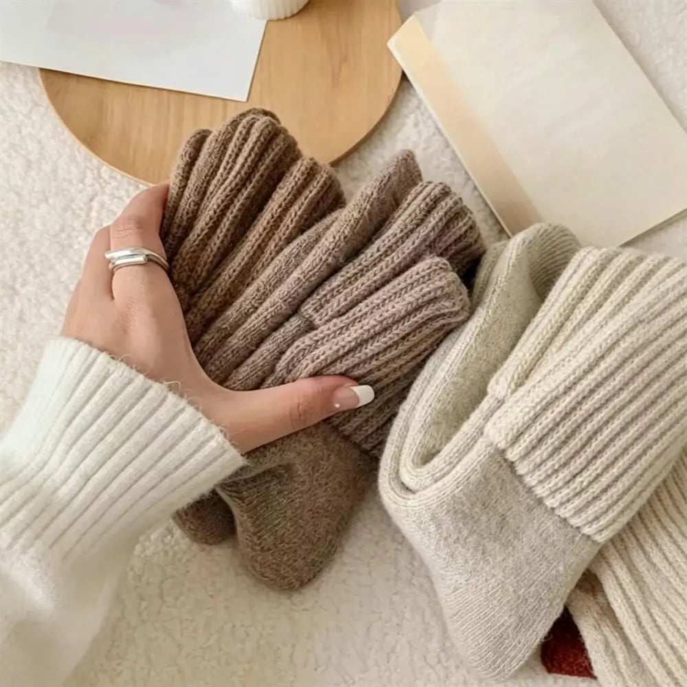 A hand wearing a silver ring holds a pair of ribbed knit socks in taupe-brown, with two more pairs in cream and taupe resting on a soft beige surface. The scene includes a wooden tray and a book as styled props.
