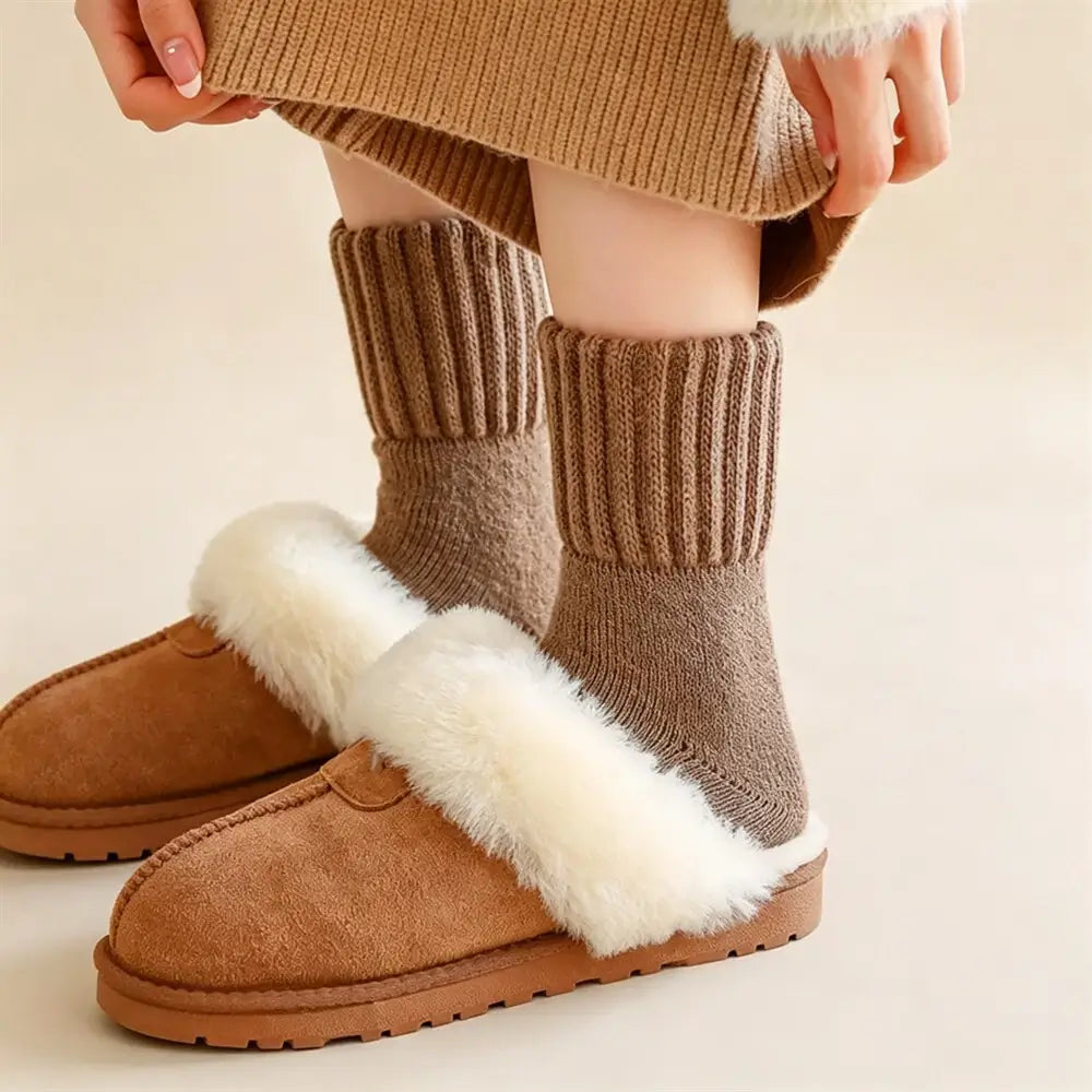 A person wearing brown knit ankle socks and tan slip-on slippers with white faux-fur trim, photographed against a soft beige studio background.