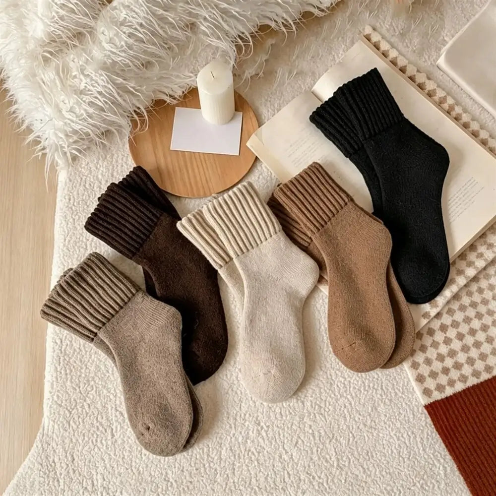 Five pairs of ribbed ankle socks in neutral tones are arranged on a textured cream rug with cozy props, including a white faux-fur throw, a candle, a small wooden tray, and an open book. The socks come in black, dark brown, taupe, cream, and camel brown.