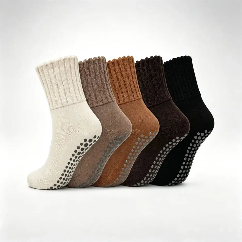 Six ankle socks arranged in a row on a light gray background, showing different color options: off-white, taupe, camel, very dark brown, charcoal, and black. Each sock has a ribbed cuff and a grip pad with raised dots on the sole.